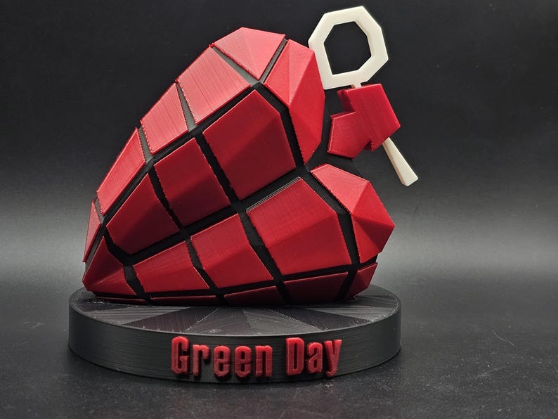 May include: A red and black 3D printed heart-shaped grenade with a white pin, on a black base with the words "Green Day" printed on it.