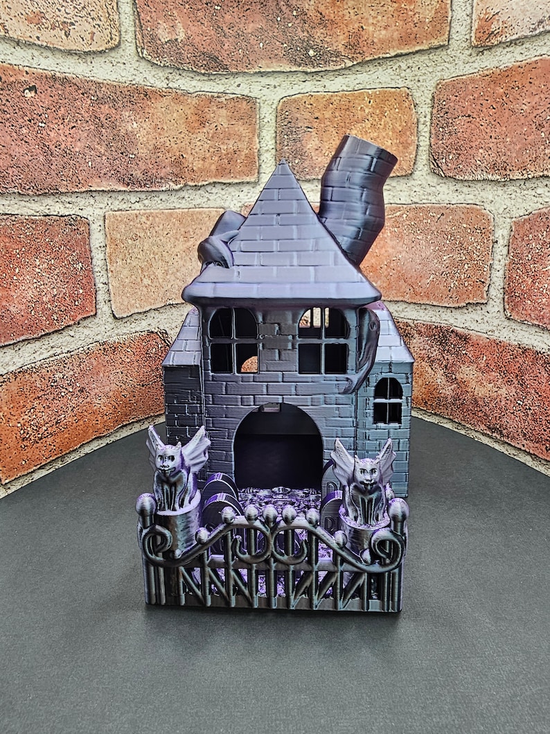 May include: A black and purple 3D printed Halloween house with a chimney, a fence, and two gargoyles. The house has a small door and windows.