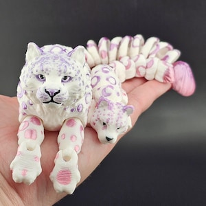 May include: A white articulated toy featuring two snow leopard heads and a long, segmented, flexible body. The figure is decorated with purple and pink spots and markings, with a smooth, matte finish and visible joints connecting the segments.