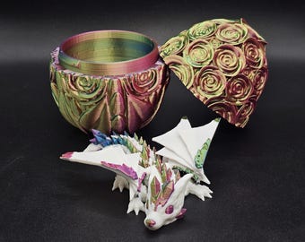 May include: A white iridescent dragon figurine with wings outstretched, sitting inside a closed iridescent egg-shaped container with a rose pattern. The egg is open and the lid is beside the dragon.