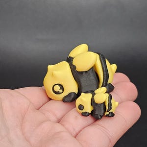 May include: A 3D printed set of two yellow and black bumblebees. The larger bee is lying on its side with its wings folded. The smaller bee is standing upright behind the larger bee.