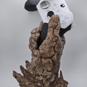 May include: A white and black Xbox controller held by a brown, textured, stone-like sculpture. The controller has black thumbsticks and buttons, with a checkered design on the centre button. The sculpture has a rough, rocky appearance.