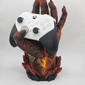 May include: A white Xbox controller with black accents rests in a detailed, red and brown monster hand-shaped stand. The stand has a textured, fiery design with a glowing orange center, creating a unique gaming accessory.