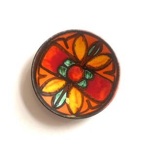 Può includere: Una piccola ciotola rotonda con un vivace motivo floreale dipinto a mano. L'interno presenta un cerchio rosso centrale circondato da petali gialli e accenti color ottanio, su uno sfondo arancione brillante con contorni marrone scuro.