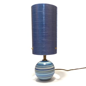 Op de afbeelding: Een tafellamp met een cilindrische blauwe textuurkap en een bolvormige voet versierd met horizontale strepen in tinten blauw, wit en groen. De lamp heeft een messingkleurige metalen hals en een dun bruin elektrisch snoer.