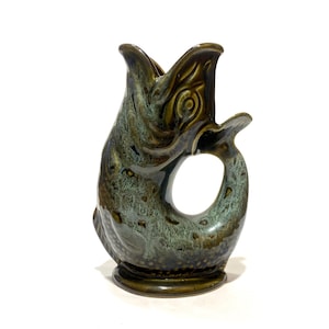 Può includere: Una brocca in ceramica a forma di pesce con la bocca aperta che funge da beccuccio. L'oggetto presenta una smaltatura maculata nei toni del verde oliva scuro e del blu ottanio, con un'apertura circolare tra il corpo e il manico a coda.