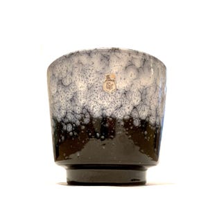 Könnte beinhalten: Eine Keramikvase mit gesprenkeltem, zweifarbigem Design. Die obere Hälfte ist hellgrau mit dunkelgrauen Sprenkeln, die in einen schwarzen Sockel übergehen. Auf der Seite ist ein kleines Etikett angebracht.