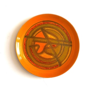 May include: A round, orange ceramic plate with a central abstract design in shades of green, brown, and orange. The plate's rim is a solid orange, and the design features intersecting lines and circular patterns.