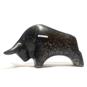 Może przedstawiać: Ciemnoszara ceramiczna figurka byka o nakrapianej fakturze. Byk przedstawiony jest w pozycji z nisko opuszczoną głową. Na boku figurki znajduje się mała, biała, prostokątna etykieta z czarnym napisem OMO KERAMIK.