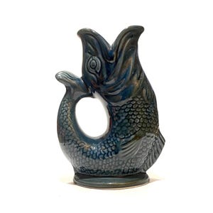 May include: A ceramic pitcher shaped like a fish with its mouth open, featuring a glossy dark blue and teal glaze. The handle is formed by the curved tail of the fish, which connects to the body, creating a circular opening in the centre.