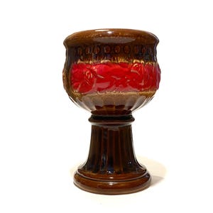 May include: A brown ceramic goblet with a pedestal base. The goblet features a band of red floral design and a rim with a series of small, raised circles. The base is a darker brown with vertical grooves.