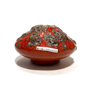 Pode incluir: Uma taça de cerâmica de perfil baixo com uma base vidrada laranja vibrante e uma superfície superior cinzento-escura texturada e com crateras. Um autocolante retangular branco com o texto preto OMO KERAMIK está colado na lateral da peça.