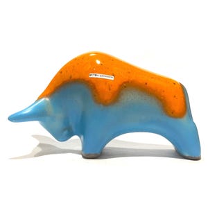 May include: A ceramic bull figurine with a glossy finish. The bull's body is light blue, and the top is orange. The figurine has a small white label that reads "OTTOKERAMIK". The bull is a decorative item.