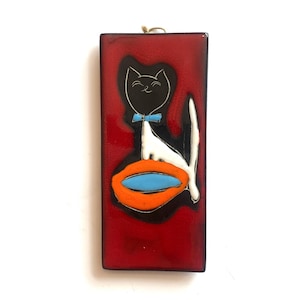 May include: A rectangular wall plaque with a glossy red finish featuring a stylised black cat with a white body, a light blue bow tie, and a long white tail. The cat sits beside a bright orange oval shape containing a light blue centre.