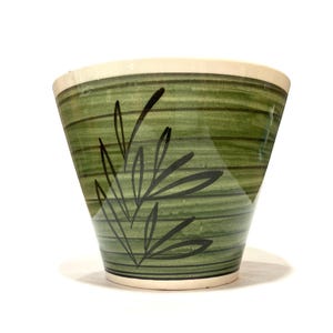 Puede incluir: Una maceta de cerámica cónica con un esmalte verde oliva brillante, rayas horizontales de color verde oscuro y un dibujo lineal negro pintado a mano de una rama con hojas en la parte frontal. El borde y la base tienen un acabado en blanco roto.
