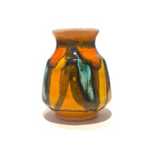 Puede incluir: Un pequeño jarrón de cerámica con cuerpo redondeado y borde acampanado. El jarrón presenta rayas verticales en tonos naranja, amarillo, negro y turquesa. La base y el borde son de color naranja liso. El jarrón está sobre un fondo blanco.