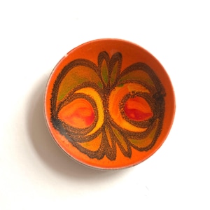 Può includere: Una ciotola rotonda e poco profonda con una base interna di colore arancione vivace. Il centro è decorato con un motivo astratto simmetrico nei toni del marrone scuro, giallo e verde, creando un effetto visivo audace e strutturato.