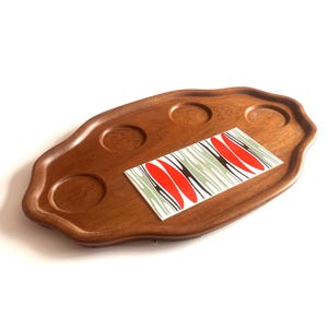 May include: An oval wooden serving tray with a scalloped edge and three circular indentations. A rectangular tile is inlaid on the surface, featuring a repeating graphic pattern of red, black, and light green abstract leaf-like shapes.