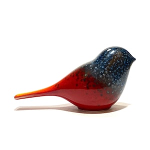 May include: A ceramic bird figurine with a glossy finish. The bird's body transitions from a vibrant red to a speckled blue and black on the head. The bird has a long, pointed tail and is perched on a white surface.