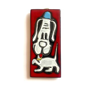 Op de afbeelding: Een rechthoekige wandplaat met een glanzende rode achtergrond en een gestileerde witte hondenillustratie. De hond heeft lange oren, een lichtblauwe hoed en een gele halsband. Het kunstwerk is uitgevoerd in een grafische stijl met zwarte omlijningen.