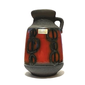 Może przedstawiać: Ceramiczny dzbanek o teksturowanej ciemnobrązowej szyjce i podstawie, z żywym pomarańczowym korpusem ozdobionym wypukłymi czarnymi kolistymi wzorami. Mała prostokątna etykieta z napisem Tonnieshof Carstens W-Germany znajduje się na przedniej stronie.