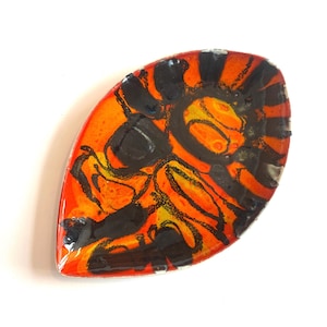 Può includere: Un piatto in ceramica a forma di foglia con una base arancione vibrante, adornato con motivi astratti neri e gialli. La superficie lucida riflette la luce, evidenziando l'audace contrasto cromatico e il design unico. Ideale per servire piccoli snack o come elemento decorativo.