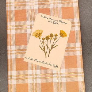 May include: A rectangular notebook with an orange and white plaid cover featuring thin gold metallic lines. Centered on the cover is a cream-colored card with a botanical illustration of yellow flowers and the text: Where Autumn Blooms into Gold, and the Heart Finds Its Light.