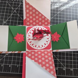 May include: A handmade Christmas-themed pop-up card. The card is red, white, and green, with snowflake and poinsettia accents. The card is open, revealing a central poinsettia arrangement and decorative cutouts.