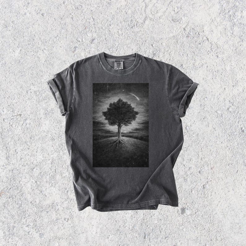 May include: Dark gray t-shirt featuring a black and white illustration of a tree with exposed roots on a hill, under a starry sky with a comet. The tee has rolled-up sleeves and a classic crew neck.