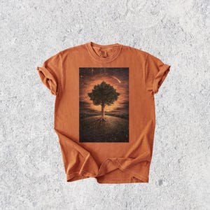 May include: An orange t-shirt featuring a rectangular graphic of a tree with exposed roots on a grassy hill, set against a sunset sky with a shooting star. The shirt has rolled-up sleeves and a crew neck.