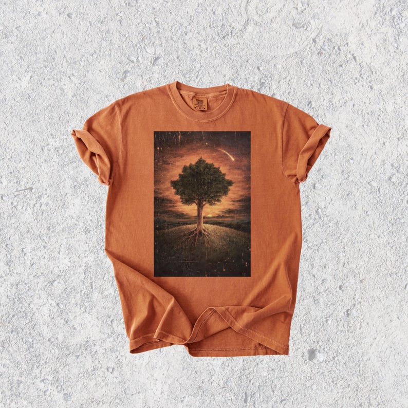 May include: An orange t-shirt featuring a rectangular graphic of a tree with exposed roots on a grassy hill, set against a sunset sky with a shooting star. The shirt has rolled-up sleeves and a crew neck.