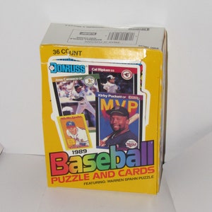 May include: A yellow box with a rainbow colored "1989 Baseball Puzzle and Cards" title. The box features a 36 count Donruss baseball card pack with a Warren Spahn puzzle. The box has images of baseball players Jose Canseco, Cal Ripken, Kirby Puckett, and a player with the Minnesota Twins.
