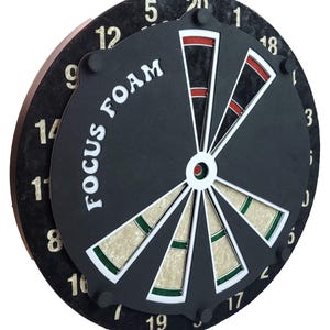 May include: A black dartboard with a white bullseye and green and white segments. The board has the words "FOCUS FOAM" printed on it in white letters. The numbers 1 through 20 are printed around the edge of the board in white.