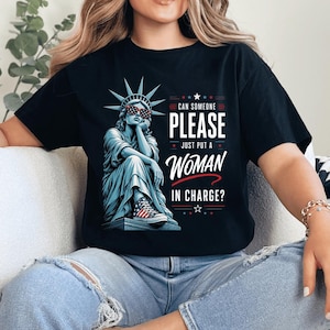 Könnte beinhalten: Ein schwarzes T-Shirt mit kurzen Ärmeln und einer Grafik der Freiheitsstatue, die eine Sonnenbrille und Turnschuhe im US-Flaggenmuster trägt. Der Text lautet: CAN SOMEONE PLEASE JUST PUT A WOMAN IN CHARGE? Das Design enthält rote Akzente.