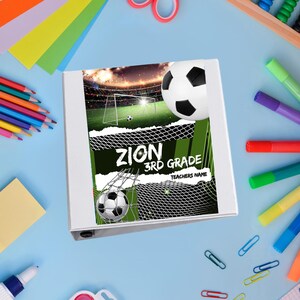 May include: White binder with a soccer-themed cover. The cover features a soccer ball, a goal, and the text "ZION 3RD GRADE TEACHERS NAME". The binder is surrounded by school supplies, including colored pencils, scissors, and paper.