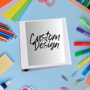 May include: A white binder with the words "Custom Design" in black script. The binder is surrounded by colorful art supplies, including colored pencils, markers, scissors, and paper. The background is light blue.