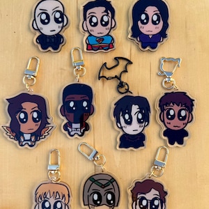 May include: A collection of cartoon character keychains with gold-colored clasps. The keychains feature various characters in a cute, stylized design. The characters are depicted in different outfits and poses, with a variety of colors used throughout the designs.