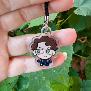 Peut inclure: Un charm en acrylique transparent représentant un personnage de dessin animé aux cheveux bruns, portant des lunettes et une tenue bleue et noire. Le charm est attaché à un cordon noir. Fond de feuillage vert flou.