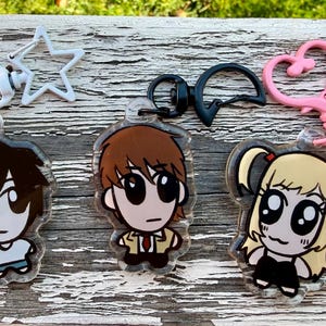 May include: Three acrylic keychains featuring anime-style characters. The keychains have different colored clasps: a white star, a black D-shaped clasp, and a pink heart-shaped clasp. The characters have black hair, brown hair, and blonde hair.