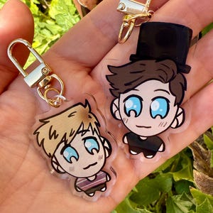 May include: Two cartoon character keychains with gold-colored clasps. One keychain features a character with blonde hair, and the other has a character with brown hair and a black top hat. Both characters have blue eyes.