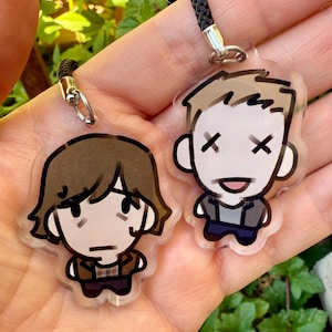 May include: Two cartoon-style acrylic keychains. One keychain features a character with brown hair and a serious expression. The other keychain has a character with light brown hair and an open mouth with X's for eyes. Both have black cords.
