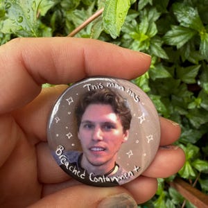 May include: A round button with a portrait of a man and the text "This man has Breached Containment". The button has a light brown background with white stars and is held in a hand against a green leafy background.