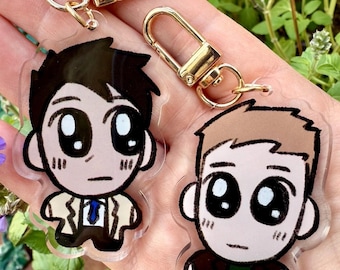 May include: Two cartoon character keychains with gold-colored clasps. One keychain features a character with black hair, a white shirt, and a blue tie. The other keychain has a character with brown hair and a dark jacket.