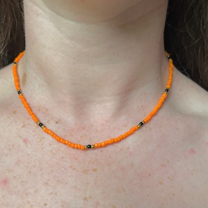 May include: A close-up of a simple orange and black beaded necklace. The necklace is made of small, round beads and is worn by a person.