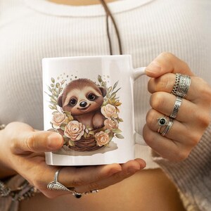 Puede incluir: Taza de cerámica blanca con un diseño de perezoso de dibujos animados rodeado de rosas rosas y vegetación. El perezoso es marrón y beige, sonriendo. La taza tiene un asa y está siendo sostenida.