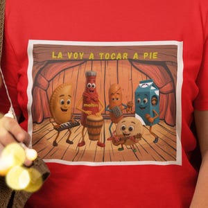Puede incluir: Camiseta roja con una imagen de dibujos animados de personajes de comida en un escenario con el texto "LA VOY A TOCAR A PIE". Los personajes incluyen una empanada tocando el piano, una botella, una zanahoria, un cartón de leche y un bollo.