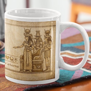 Puede incluir: Taza de cerámica blanca con una ilustración del antiguo Egipto en tonos sepia. El diseño incluye figuras, jeroglíficos y la palabra "eterno". La taza está sobre una mesa con galletas y una jarra de metal.