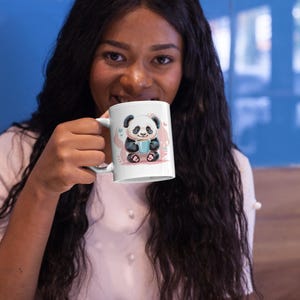 Puede incluir: Taza de cerámica blanca con un diseño de oso panda. El panda sostiene una taza azul, rodeado de corazones y flores rosas. La taza tiene un asa blanca. La taza es sostenida por una persona.