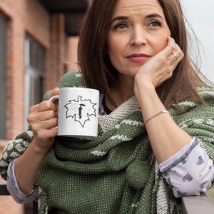 Puede incluir: Taza de cerámica blanca con un dibujo lineal negro de una persona con un paraguas dentro de un contorno de hoja. La persona lleva un chal verde con un estampado blanco. El fondo incluye un edificio de ladrillo y un cielo nublado.
