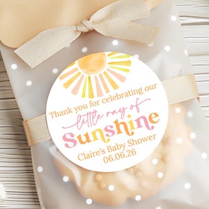May include: A baby shower favor bag with a cookie inside. The bag is clear with white polka dots and tied with a beige ribbon. A round sticker reads "Thank you for celebrating our little ray of Sunshine" with a sun graphic.